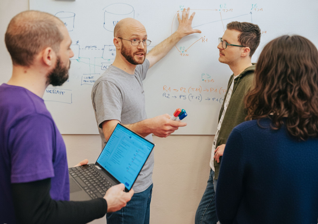 Quattro ragazzi discutono davanti a una whiteboard. Uno ha un computer in mano, uno sta gesticolando alla lavagna e due stanno ascoltando attenti.