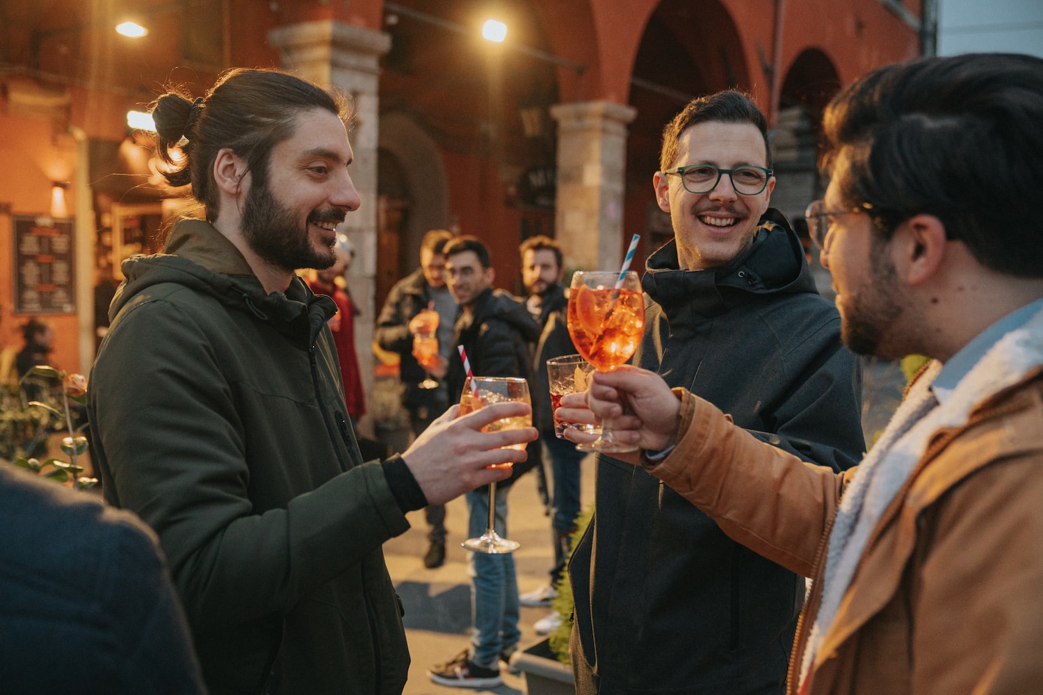 Tre ragazzi che fanno un brindisi con dei bicchieri di spritz in una piazza di Pisa di sera
