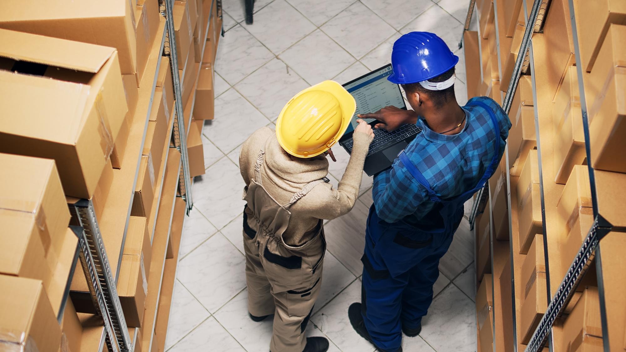 Due operatori logistica che discutono lavorando a un computer portatile tra due file di scaffali di magazzino